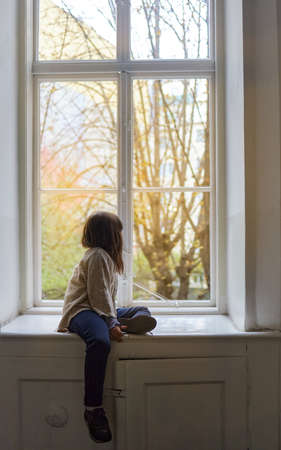 lonely girl sitting on the side of a large antique window looking out onto the street.の写真素材