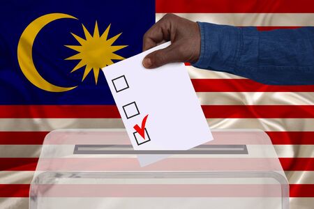 male voter drops a ballot in a transparent ballot box against the Malaysia national flag, concept of...