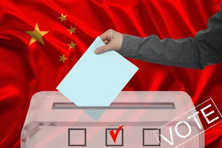 male voter drops a ballot in a transparent ballot box against the background of the national flag of...