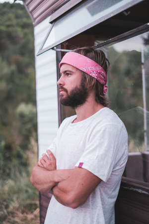 Young man with a long hair and bread, white shirt and pink scarf looking to camera near a mobile home in the woodsの写真素材