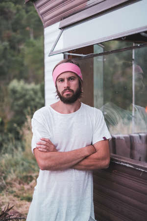 Young man with a long hair and bread, white shirt and pink scarf looking to camera near a mobile home in the woodsの写真素材