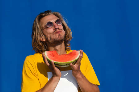 Attractive young man with long blond hair with half a watermelon covering his face on a plain blue background. Summer, sun, heat, fruit, vacation concept.の写真素材
