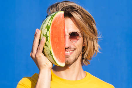 Attractive young man with long blond hair with half a watermelon covering his face on a plain blue background. Summer, sun, heat, fruit, vacation concept.の写真素材