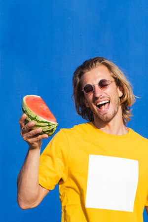 Attractive young man with long blond hair with half a watermelon covering his face on a plain blue background. Summer, sun, heat, fruit, vacation concept.の写真素材