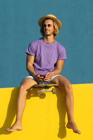 Blond young man with skateboard, mobile phone, hat and sunglasses enjoying the summer. Taking a...