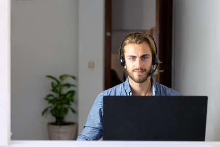 Blond male working from home with headphones using wireless internet on laptop, man call center agent or telemarketerの写真素材