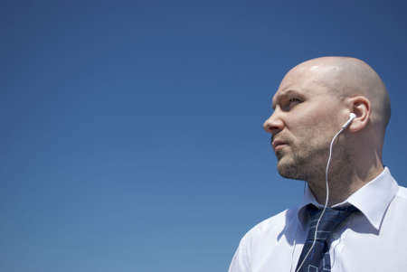 Royalty free stock image of an unshaven office worker wearing white headphones looking out to distance against clean blue sky providing copy space to the left, vignettedの写真素材
