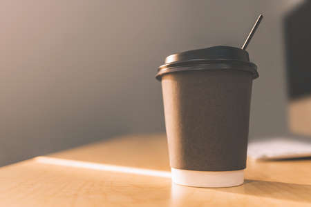 take away paper cup of hot coffee with computer keyboard on top of wooden working table in morning sunlight from window and selective focusingの写真素材