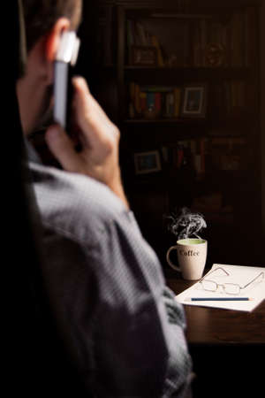 Smoky coffee cup, a pencil and glasses in the foreground, with a man speaking by phone sitting in a...