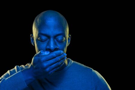 Colorful portrait of a black man with mouth covered by hand and eyes closed. Vibrant light with saturated blue gel filter and black background. Horizontal with copyspaceの写真素材
