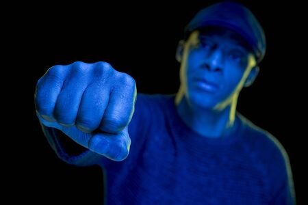 Close-up of a fist. Black man with blue and yellow light, wearing a flat cap, isolated on black background, looking at the camera. Horizontalの写真素材