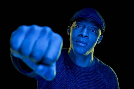 Black man with blue and yellow light, wearing a flat cap, isolated on black background, looking at the camera. Unfocused fist in foreground. Horizontalの写真素材