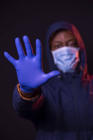 STOP Coronavirus, African American man shows the stop sign, wearing protective mask and gloves on dark background. Focus on hand. COVID-19 concept.の写真素材