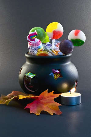 Against a dark background, a dark china cauldron is bursting with candies and lollipops.  Silk fall leaves and candle in the foreground.の写真素材