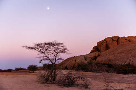 A bright full moon accentuates a red glowing desert and a pink sky. Namibian desert near Spitzkoppe.の写真素材