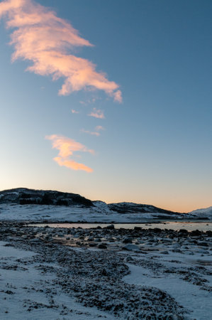 A bright orange glow colours the golden hour sky that serves as a dramatic background for the rugged mountains and snow-covered beaches of arctic norway, near the town of Bodo.の写真素材