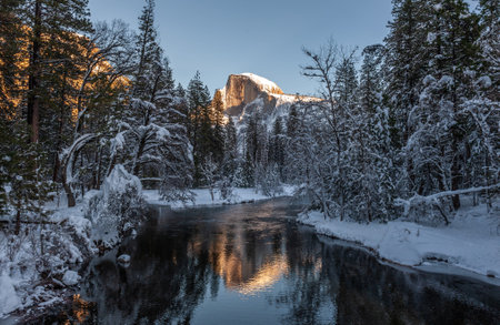 The peak of half dome is baking in the full light of the setting sun. It is reflected in the merced river, on a bright snowy winter afternoon during golden hour.の写真素材