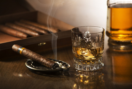 Glass of whiskey with ice cubes and smoking cigar on wooden tableの写真素材