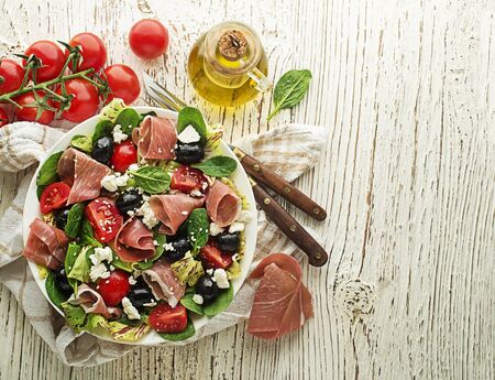 Healthy green salad with prosciutto, olives, tomato and fresh cheese on wooden table background. Healthy meal.の写真素材
