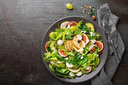 Fresh green leafy salad with mozzarella cheese, fruits and nuts on dark background close up. Concept for a tasty and healthy mealの写真素材