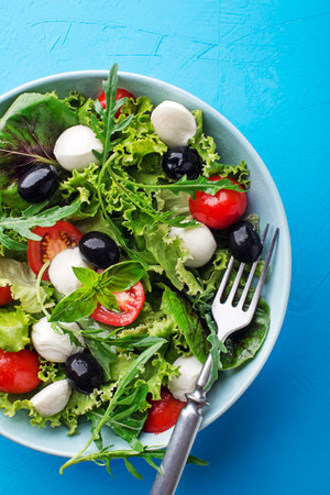 Caprese Italian or Mediterranean salad. Green Salad with mozzarella, black olives and tomato on blue table background close upの写真素材