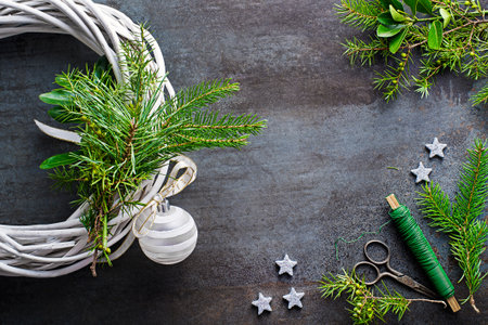 Making christmas wreath from natural branches with white bauble ball, ribbon bow and shiny stars on dark grey backgroundの写真素材