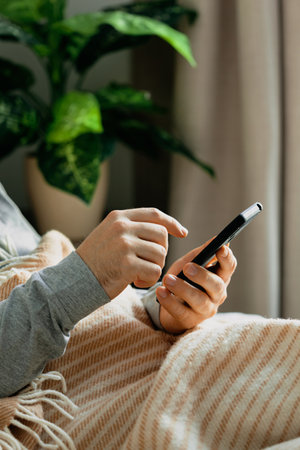 a caucasian man relaxing, using smart phone lying in bed under throw at homeの写真素材