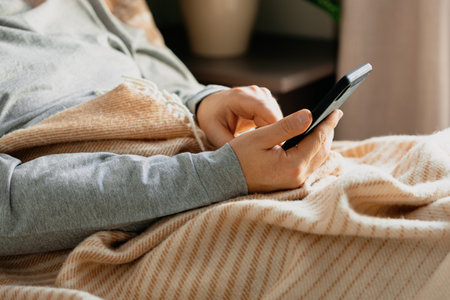 a caucasian man relaxing, using smart phone lying in bed under throw at homeの写真素材