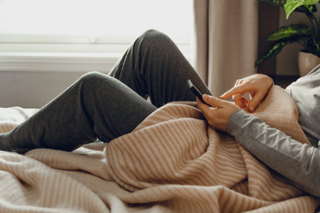 a caucasian man relaxing, using smart phone lying in bed under throw at homeの写真素材
