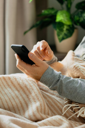 a caucasian man relaxing, using smart phone lying in bed under throw at homeの写真素材