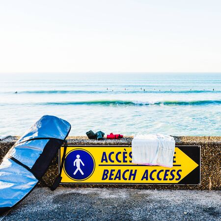Surfboard in its bag next to a beach access sign, ocean in the background with silhouettes of surfers, in Bidart, France.の写真素材