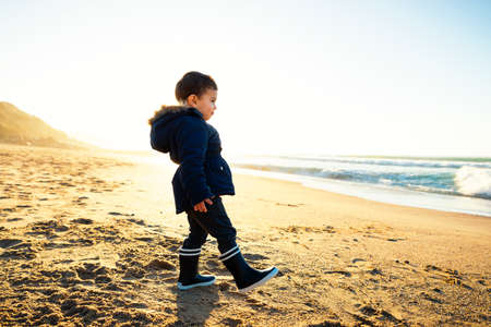 Backlit cute little boy walking on beach at sunset, winter season. Toddler wearing dark blue coat and gumboots.の写真素材