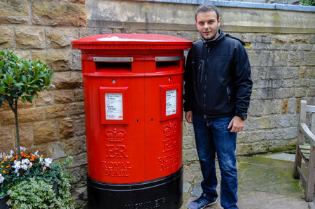 Man standing next to a bright red Royal Mail post box in Windsor, England, a traditional British...
