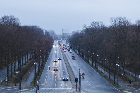 Rainy evening view from the Victory Column in Berlin, Germany. Cars and a motorbike move along a wet boulevard lined with bare trees, with city buildings in the distance.の写真素材