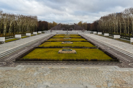 Symmetrical gardens at Soviet War Memorial in Treptower Park, Berlin, with lawns, paved paths, and monumental stone walls under cloudy sky.の写真素材
