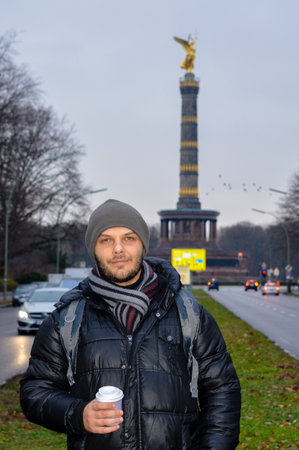 Portrait of a young man in winter clothing holding a coffee cup, standing on the road with the Berlin Victory Column (SiegessÃ¤ule) in the background, Berlin, Germany.の写真素材