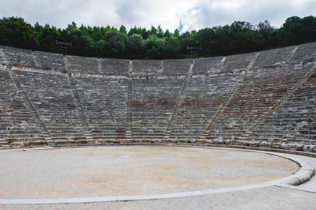 Wide view of the Ancient Epidaurus Theater in Greece, with its stone seating rows, central circular stage and surrounding green trees, a famous UNESCO archaeological landmark.の写真素材