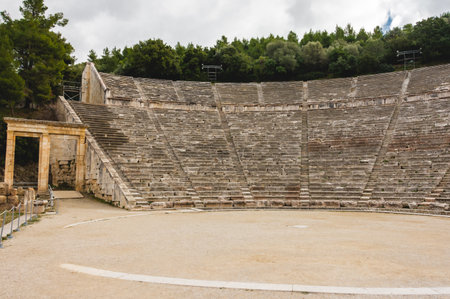 Wide angle view of the Ancient Epidaurus Theater in Greece, with its historic stone seating rows and preserved entrance gate on the left side, framed by green trees and sky.の写真素材