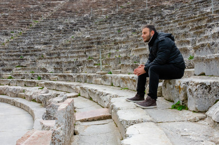 Man in black jacket sitting on stone seats of the Ancient Epidaurus Theater in Greece, looking aside.の写真素材