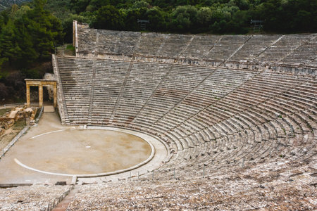 Panoramic view from the top of the Ancient Epidaurus Theater in Greece, with its stone seating rows, circular stage and preserved entrance gate, surrounded by lush green trees.の写真素材