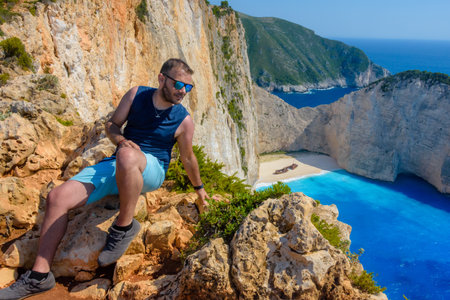 Traveler sitting on cliff edge overlooking Navagio shipwreck beach in Zakynthos, Greece. A vibrant travel portrait with iconic view, turquoise sea, and summer vibes.の写真素材