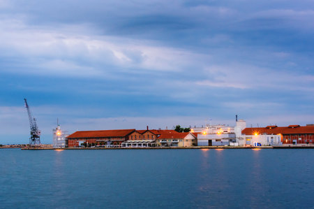 Industrial buildings and port warehouses at Thessaloniki, Greece, photographed at twilight with lights glowing and dramatic clouds above the calm blue sea.の写真素材