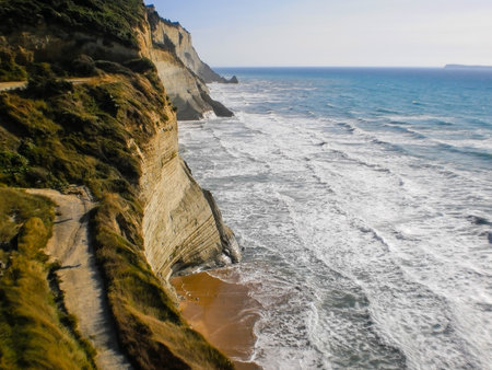 Aerial view of the steep cliffs above Loggas Beach in Peroulades, Corfu, Greece. Crashing waves, golden sand, and a scenic path leading along the dramatic coastline.の写真素材