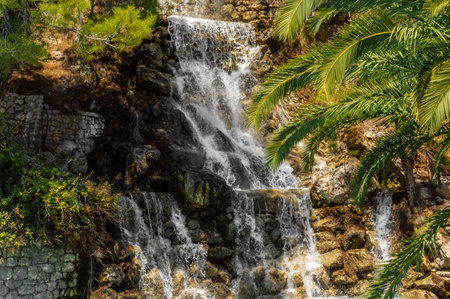 A vibrant waterfall cascades over rocky stones surrounded by lush greenery and palm trees in Loutraki, Corinth, Greece. A peaceful summer scene in bright sunlight.の写真素材