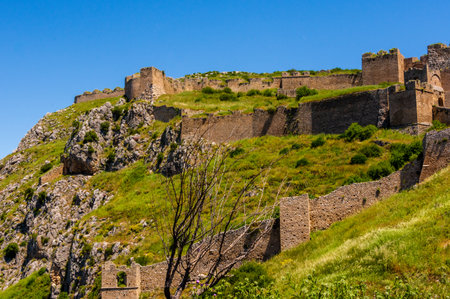 Historic fortress walls of Acrocorinth stretch across the rugged green hillside under a vivid blue sky in the Peloponnese region of southern Greece.の写真素材