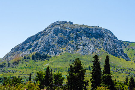 The ancient fortress ruins of Acrocorinth sit atop a dramatic rocky mountain in Ancient Corinth, Greece, with green vegetation and bright clear sky creating a scenic view.の写真素材