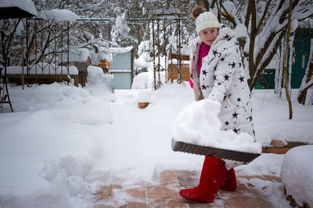 A woman with a shovel clears snow from the tile area. The girl cleans the snow in the yard. Snow cover on the knee. in red felt bootsの写真素材