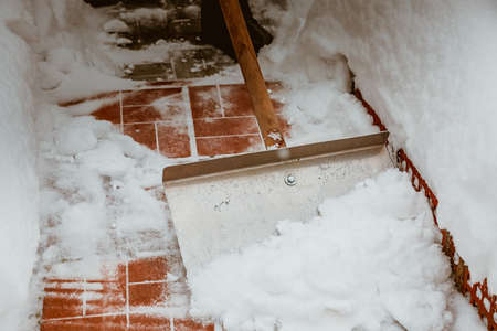 Snow removal with an iron shovel. With a wooden handle. Winter cleaning of the yard from the snow. After a heavy snowfall. Cleaning the steps from red tilesの写真素材
