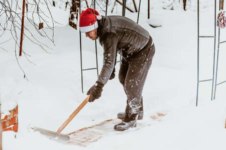 A man cleans steps from snow. In a red hat of Santa Claus and a leather black jacket. In felt boots. Clearing snow from the backyard after a snowfall.の写真素材