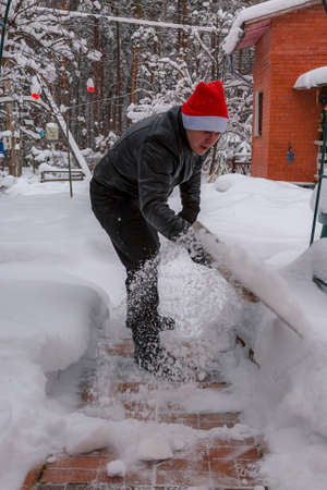 A man cleans steps from snow. In a red hat of Santa Claus and a leather black jacket. In felt boots. Clearing snow from the backyard after a snowfall.の写真素材